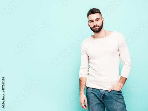 Portrait of handsome confident stylish hipster lambersexual model.Man dressed in white sweater and jeans. Fashion male isolated on blue wall in studio. Thoughtful