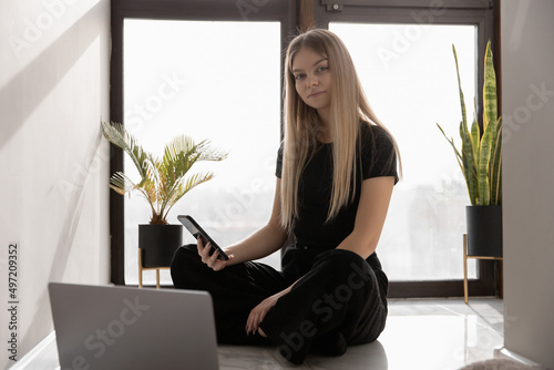 beautiful woman works at home at the computer sitting on the floor by the window and looking smiling at the camera with a teleon in her hand photo