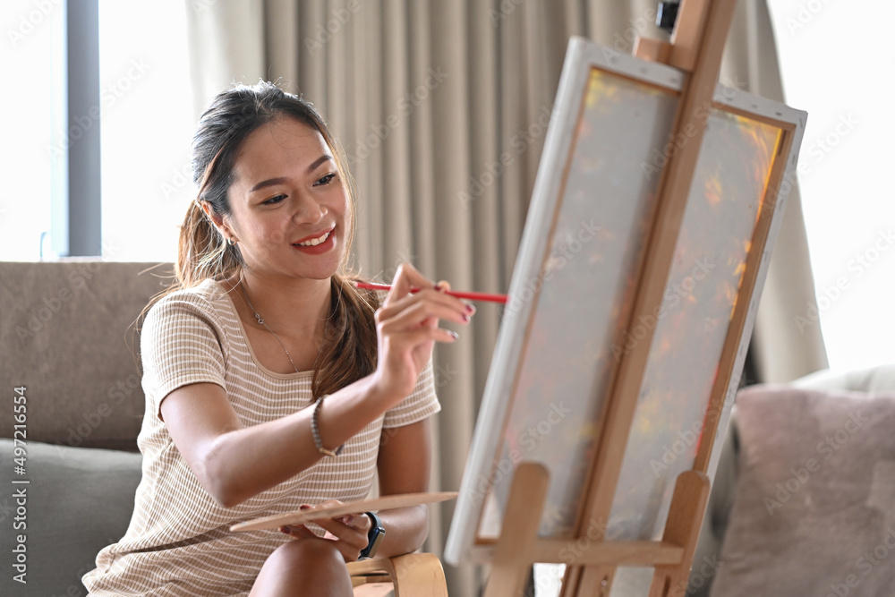 Charming young woman sitting in living room and painting picture with acrylic paint.