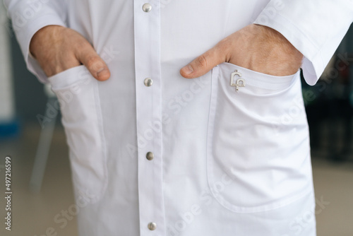 Close-up cropped shot of unrecognizable male doctor wearing white medical uniform holding hands in pockets standing in hospital office. Practitioner man posing at workplace. Concept of medical work.