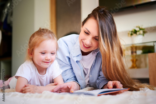 Young mother with her cute little daughter is reading book and looking at the drawings in the book while lying on the floor. The mother teaches the child. The relationship between parents and children