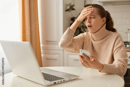 Indoor shot of astonished scared woman wearing beige sweater posing in kitchen, looking at cell phone screen with open mouth, touching head, forgot important information, mistake.