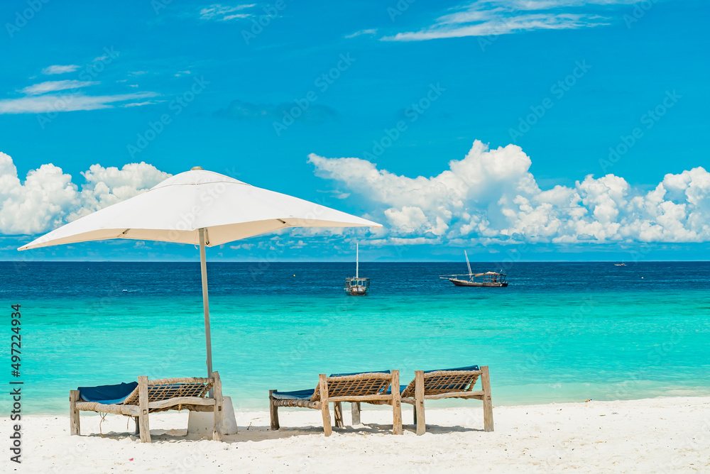 Obraz premium Wooden deck chairs on a sandy beach in front of ocean with boats, Zanzibar Tanzania