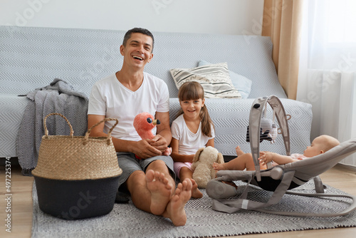 Portrait of happy satisfied man wearing white casual T-shirt sitting on floor with his daughters, smiling man with elder child looking at camera, playing together with infant baby in rocking chair.