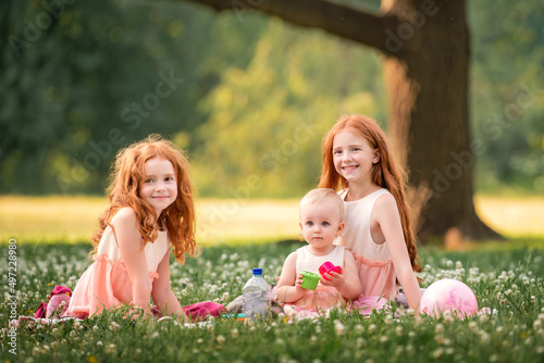 Fotografia Three red-haired curly happy sisters children play picnic on the grass among whi