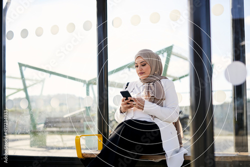 Woman in headscarf waiting at bus stop and using phone photo