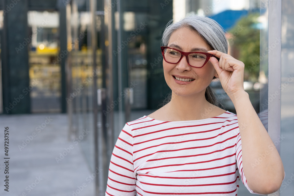 Confident mature businesswoman wearing red stylish eyeglasses standing on urban street, copy space. Portrait of smiling asian manager looking away outdoors. Successful business Confident mature businesswoman wearing red stylish eyeglasses standing on urban street, copy space. Portrait of smiling asian manager looking away outdoors. Successful business