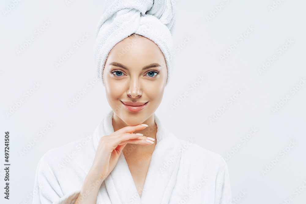 Woman looks with calm face, enjoys bath procedures, wears soft white robe, wrapped towel on head, isolated on white background Woman looks with calm face, enjoys bath procedures, wears soft white robe, wrapped towel on head, isolated on white background