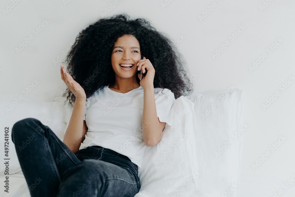 Afro American woman has telephone talk, gestures and laughs with joy, looks somewhere aside, spends time at home, sits on white bed Afro American woman has telephone talk, gestures and laughs with joy, looks somewhere aside, spends time at home, sits on white bed
