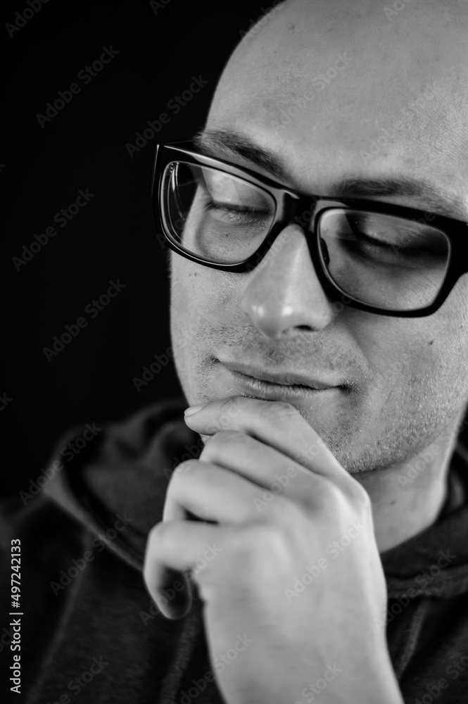 Close up portrait in black and white of a young bald man in black eyeglasses holding his chin with his hand while thinking looking down Close up portrait in black and white of a young bald man in black eyeglasses holding his chin with his hand while thinking looking down