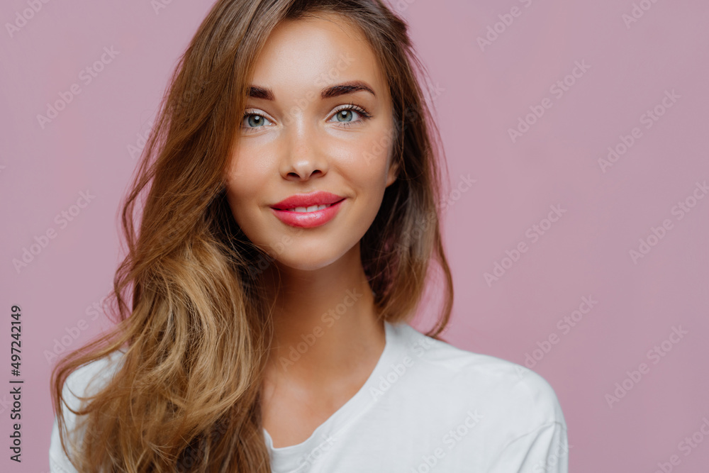 Female model has tender smile, wears minimal makeup, has long wavy hair, looks at camera with satisfaction Female model has tender smile, wears minimal makeup, has long wavy hair, looks at camera with satisfaction