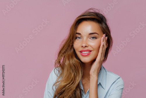 Female model touches cheeks with palm, has gentle toothy smile, wears makeup, stands against purple wall, copy space area for your advertisement