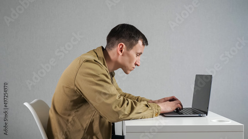 Canvas Print Young man works on laptop sitting at white table in office against grey wall