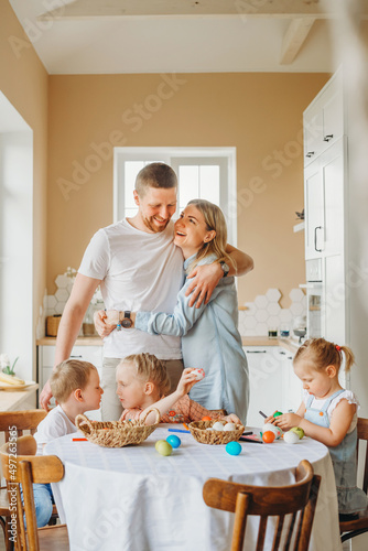 a happy family with three children is preparing for Easter in the kitchen at home, parents hug and smile, and children decorate colorful Easter eggs