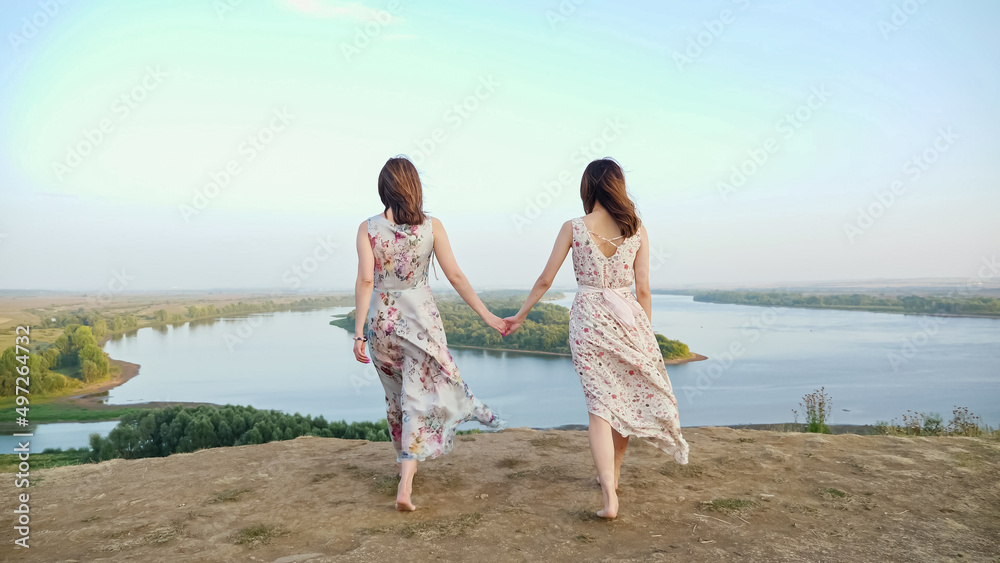 Graceful brunette barefoot women in long dresses walk joining hands along hilly riverbank at countryside on windy summer day backside view Graceful brunette barefoot women in long dresses walk joining hands along hilly riverbank at countryside on windy summer day backside view