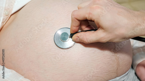 hand of an unrecognizable man drives a stethoscope over the big belly of a pregnant woman.