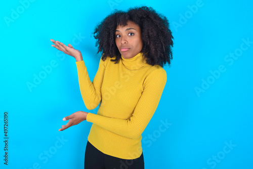 Young woman with afro hairstyle wearing yellow turtleneck over blue background pointing aside with both hands showing something strange and saying: I don't know what is this. Advertisement concept.