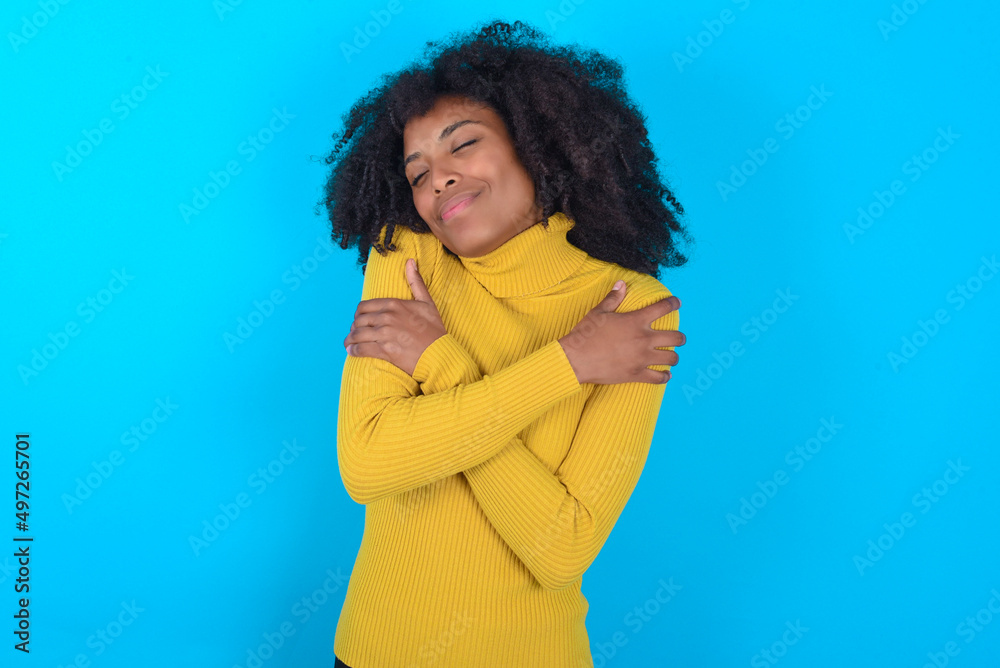 Young woman with afro hairstyle wearing yellow turtleneck over blue background Hugging oneself happy and positive, smiling confident. Self love and self care Young woman with afro hairstyle wearing yellow turtleneck over blue background Hugging oneself happy and positive, smiling confident. Self love and self care