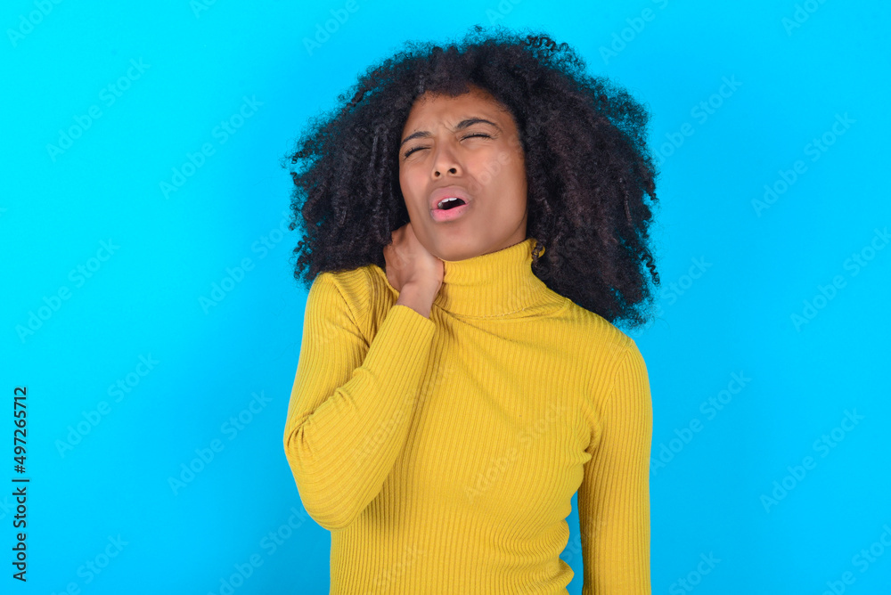 Young woman with afro hairstyle wearing yellow turtleneck over blue background Suffering of neck ache injury, touching neck with hand, muscular pain Young woman with afro hairstyle wearing yellow turtleneck over blue background Suffering of neck ache injury, touching neck with hand, muscular pain