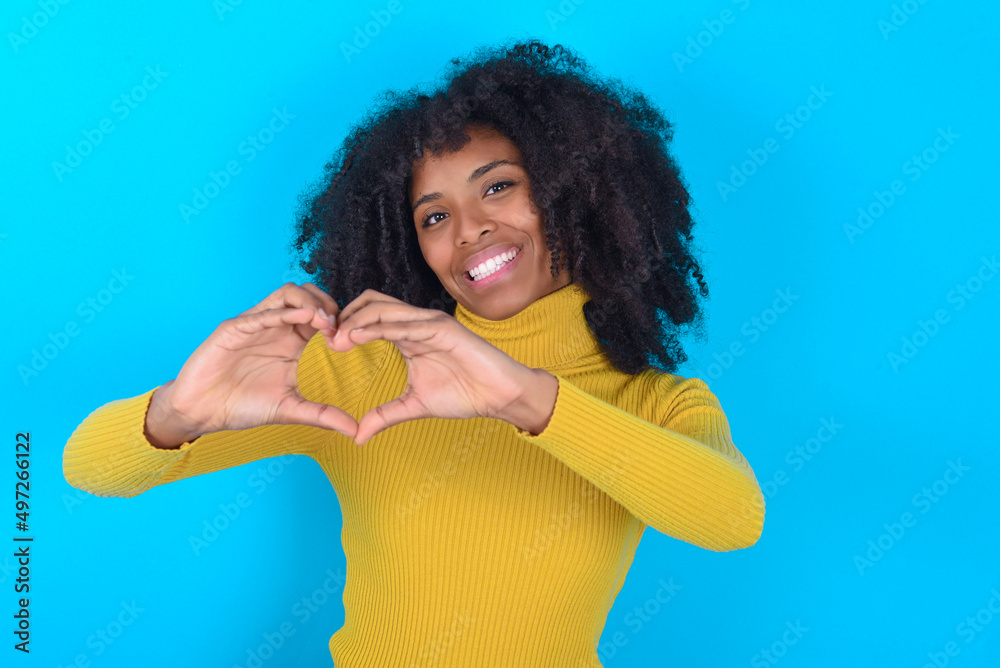 Young woman with afro hairstyle wearing yellow turtleneck over blue background smiling in love doing heart symbol shape with hands. Romantic concept. Young woman with afro hairstyle wearing yellow turtleneck over blue background smiling in love doing heart symbol shape with hands. Romantic concept.