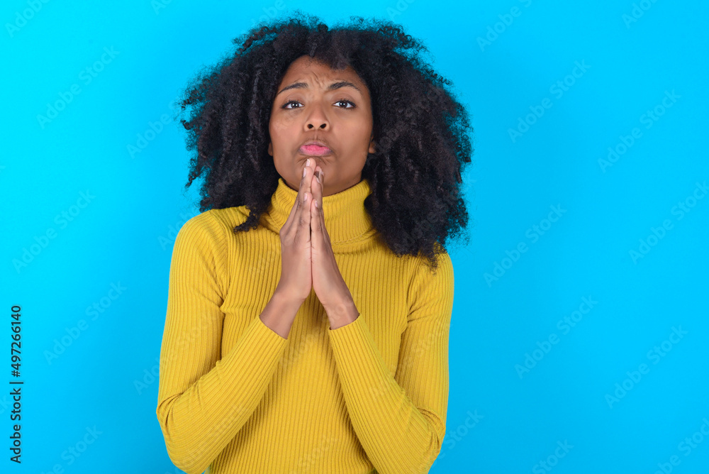 Positive Young woman with afro hairstyle wearing yellow turtleneck over blue background smiles happily, glad to receive pleasant news from interlocutor, keeps palms together, People emotions concept. Positive Young woman with afro hairstyle wearing yellow turtleneck over blue background smiles happily, glad to receive pleasant news from interlocutor, keeps palms together, People emotions concept.