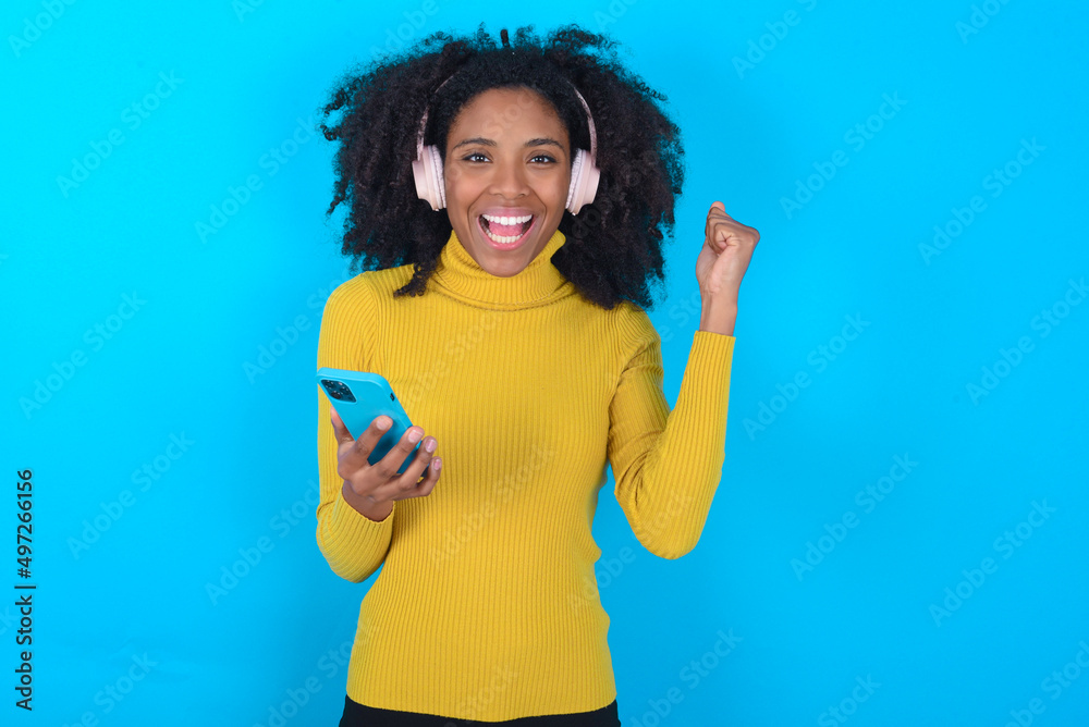 Positive Young woman with afro hairstyle wearing yellow turtleneck over blue background holds modern cell phone connected to headphones, clenches fist from good emotions, exclaims with joy, Positive Young woman with afro hairstyle wearing yellow turtleneck over blue background holds modern cell phone connected to headphones, clenches fist from good emotions, exclaims with joy,