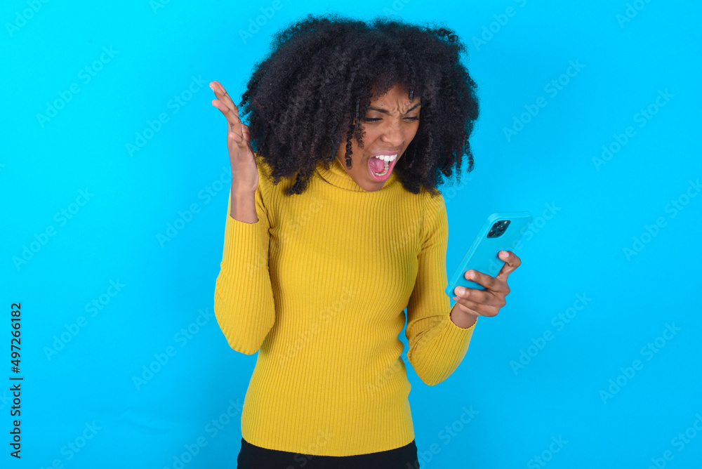 Angry Young woman with afro hairstyle wearing yellow turtleneck over blue background screaming on the phone, having an argument with an employee. Troubles at work. Angry Young woman with afro hairstyle wearing yellow turtleneck over blue background screaming on the phone, having an argument with an employee. Troubles at work.