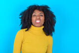 Young woman with afro hairstyle wearing yellow turtleneck over blue background keeps teeth clenched, frowns face in dissatisfaction, irritated because of much duties.