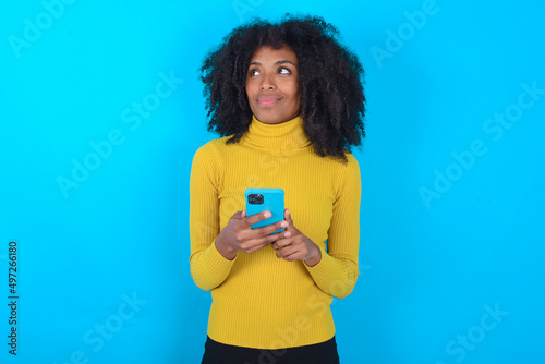 Young woman with afro hairstyle wearing yellow turtleneck over blue background holds telephone hands reads good youth news look empty space advert