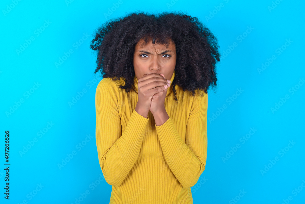 Young woman with afro hairstyle wearing yellow turtleneck over blue background holding oneself, feels very cold outside, hopes that will not get cold Young woman with afro hairstyle wearing yellow turtleneck over blue background holding oneself, feels very cold outside, hopes that will not get cold