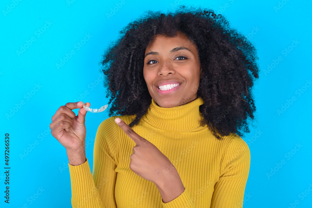 Young woman with afro hairstyle wearing yellow turtleneck over blue background holding an invisible aligner and pointing at it. Dental healthcare and confidence concept. Young woman with afro hairstyle wearing yellow turtleneck over blue background holding an invisible aligner and pointing at it. Dental healthcare and confidence concept.