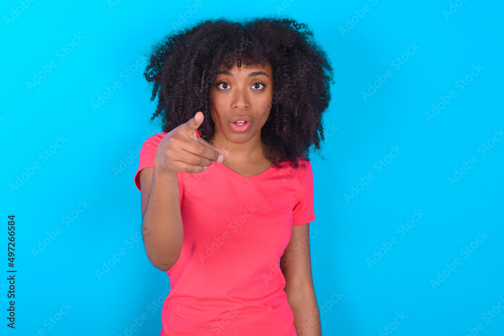 Shocked Young girl with afro hairstyle wearing pink t-shirt over blue background points at you with stunned expression Shocked Young girl with afro hairstyle wearing pink t-shirt over blue background points at you with stunned expression