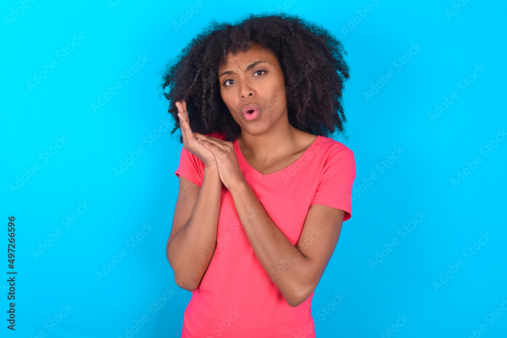 Surprised emotional Young girl with afro hairstyle wearing pink t-shirt over blue background rubs palms and stares at camera with disbelief Surprised emotional Young girl with afro hairstyle wearing pink t-shirt over blue background rubs palms and stares at camera with disbelief