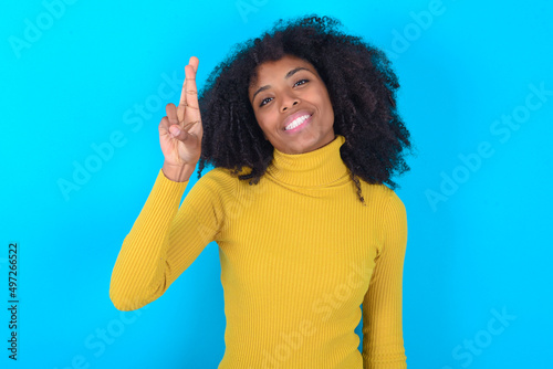 Young woman with afro hairstyle wearing yellow turtleneck over blue background pointing up with fingers number ten in Chinese sign language Shi photo