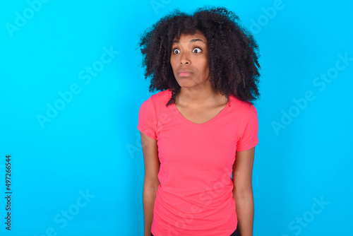 Young girl with afro hairstyle wearing pink t-shirt over blue background stares aside with wondered expression has speechless expression. Embarrassed model looks in surprise