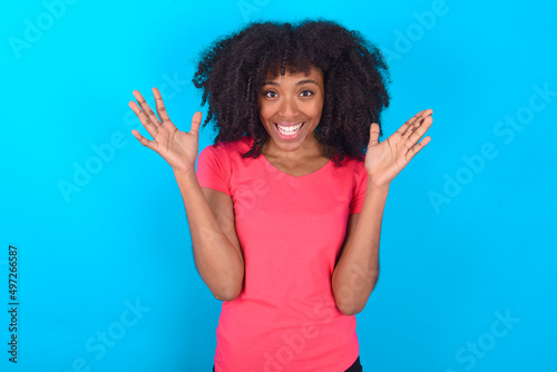 Crazy outraged Young girl with afro hairstyle wearing pink t-shirt over blue background screams loudly and gestures angrily yells furiously. Negative human emotions feelings concept