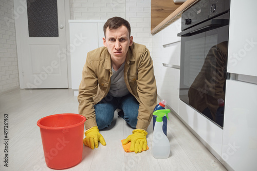 Man cleans laminate floor with detergent and water in plastic bucket smiling in modern kitchen. Young male housekeeper takes care of apartment doing chores, closeup