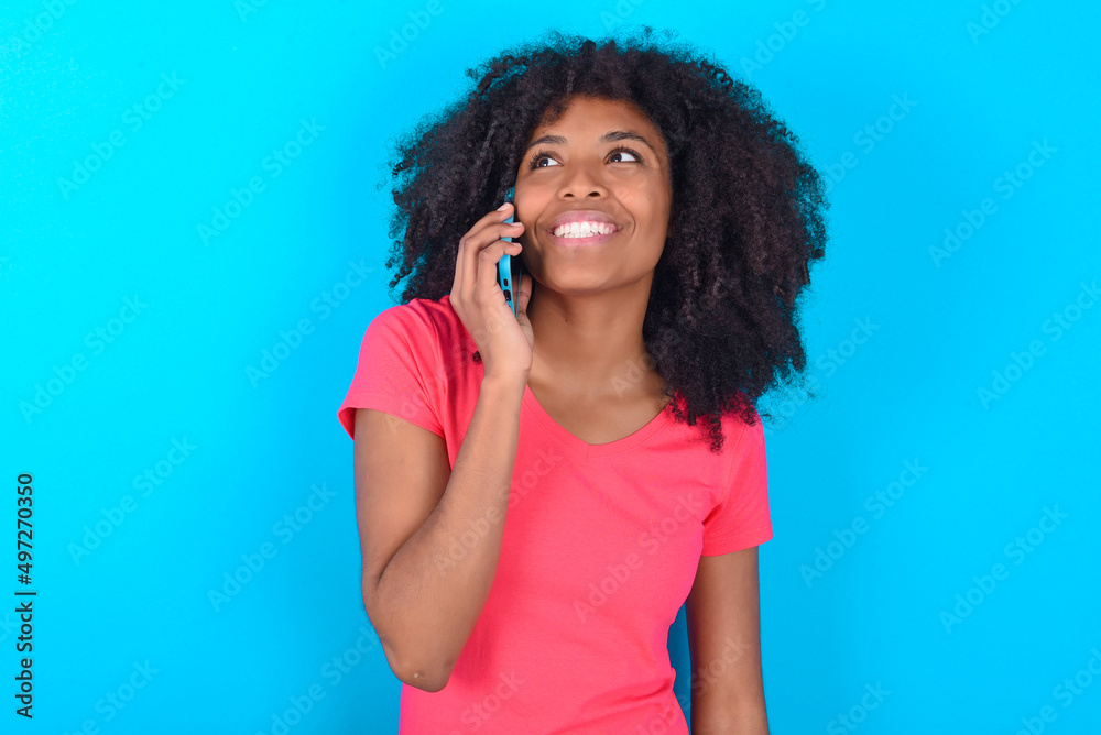 Pleasant looking happy Young girl with afro hairstyle wearing pink t-shirt over blue has nice telephone conversation and looks aside, has nice mood and smiles positively while talks via cell phone Pleasant looking happy Young girl with afro hairstyle wearing pink t-shirt over blue has nice telephone conversation and looks aside, has nice mood and smiles positively while talks via cell phone