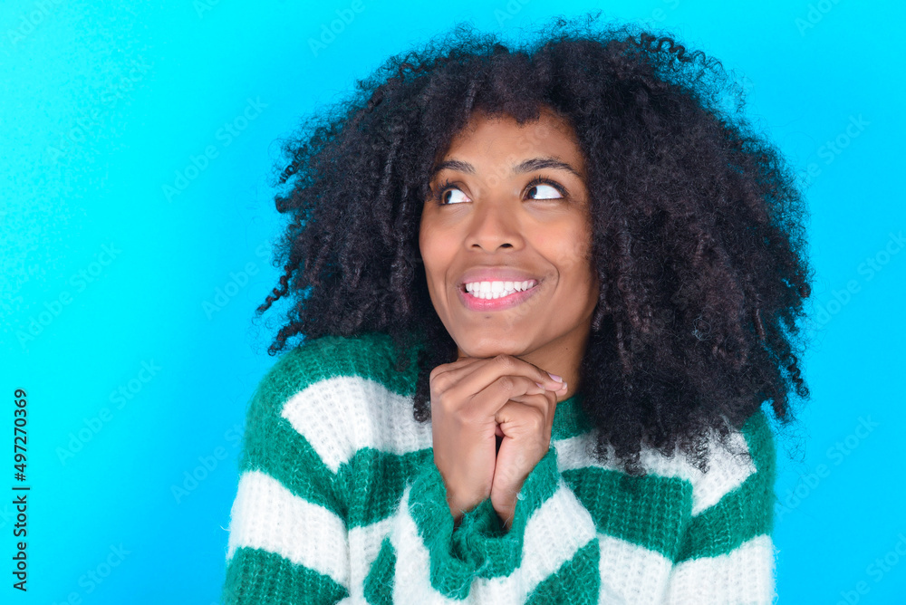 Happy Young woman with afro hairstyle wearing striped sweater over blue background anticipates something awesome happen, looks happily aside, keeps hands together near face, has glad expression. Happy Young woman with afro hairstyle wearing striped sweater over blue background anticipates something awesome happen, looks happily aside, keeps hands together near face, has glad expression.