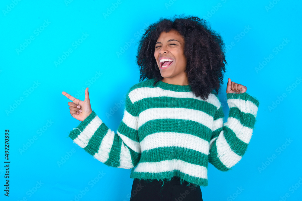 Cheerful Young woman with afro hairstyle wearing striped sweater over blue background showing copy space ad celebrating luck Cheerful Young woman with afro hairstyle wearing striped sweater over blue background showing copy space ad celebrating luck