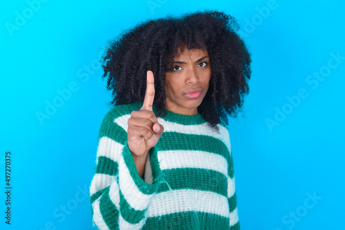 Young woman with afro hairstyle wearing striped sweater over blue background frustrated and pointing to the front