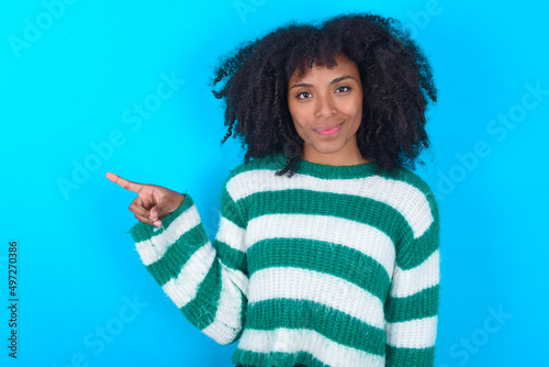 Young woman with afro hairstyle wearing striped sweater over blue background points to side on blank space demonstrates advertisement. People and promotion concept © Roquillo