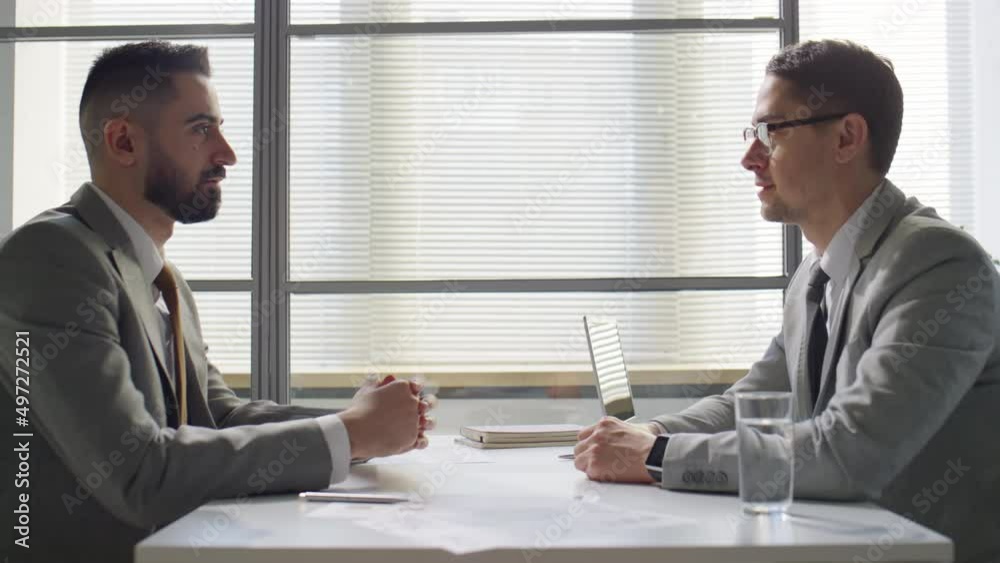 Side view of middle eastern and caucasian businessmen having discussion and shaking hands at table in office Side view of middle eastern and caucasian businessmen having discussion and shaking hands at table in office