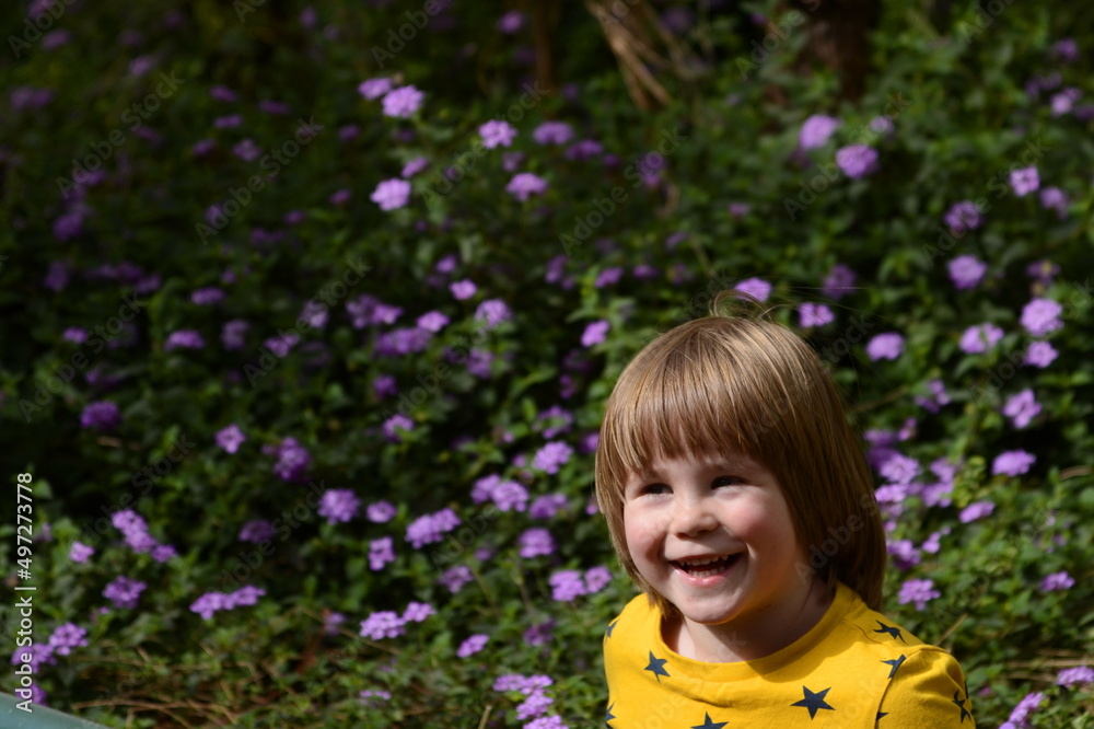 Funny baby in a yellow jacket on a background of purple flowers. Little boy, three years old. Blond hair, sweet smile Funny baby in a yellow jacket on a background of purple flowers. Little boy, three years old. Blond hair, sweet smile