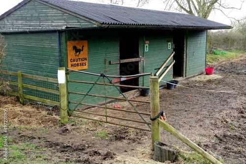 Horse stable in muddy field behind metal gate.