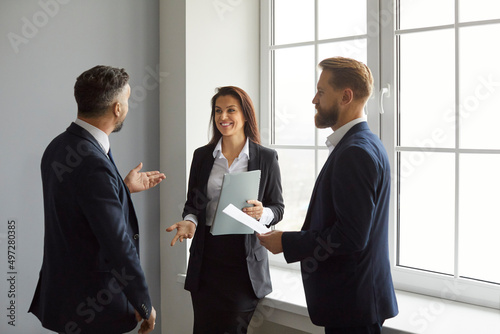 Business professionals. Smiling business people discuss work, project strategy and exchange ideas together. Two men and woman in business clothes stand with documents in hand near window in office.