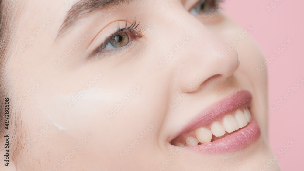 Extreme close-up of young good-looking slim European skincare woman with cream smear under her eye smiling wide against pink background | Eye cream commercial concept