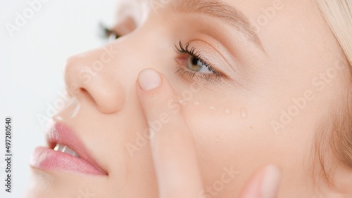 Extreme close-up of young Caucasian woman with pure skin applies face serum smearing drops on her cheek on white background | Skin care serum commercial concept