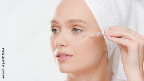 Big close-up shot of young Caucasian woman with hair-drying towel on her head applies face serum on her cheek from dropper on white background
