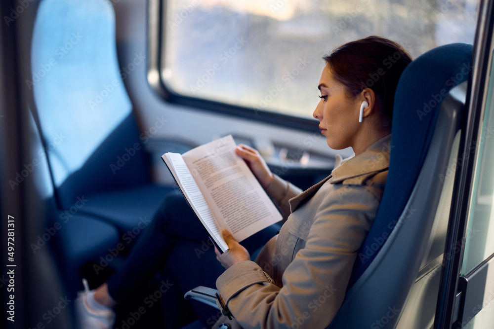 Young woman reads novel while listening music over earbuds in train. - obrazy, fototapety, plakaty 